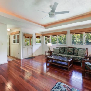 A spacious living room with hardwood floors and large windows, offering a view of the lush greenery outside. Ideal for a tranquil stay in Kauai vacation rentals. Spacious living room with hardwood floors and large windows overlooking greenery.